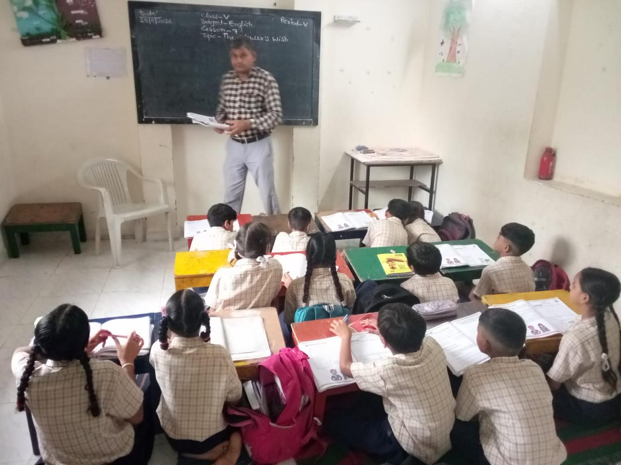 A teacher reads to Class V from the front of the room during an English lesson, pupils following along in their workbooks at low desks.