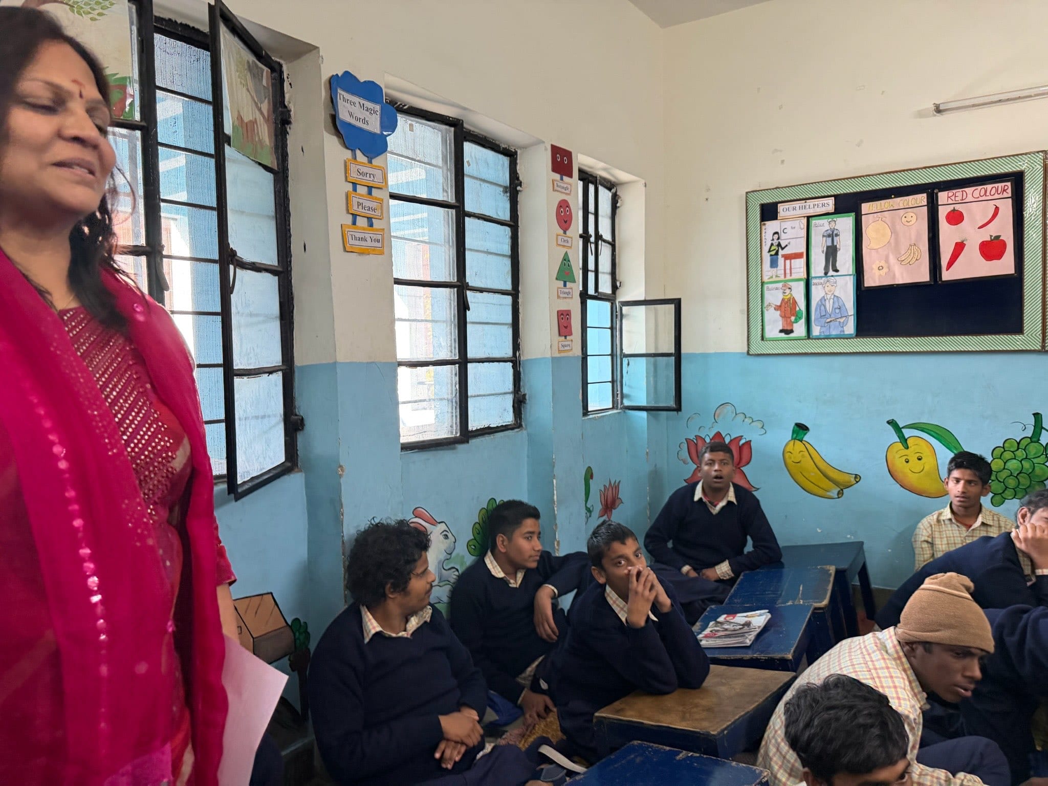 A visitor in a bright pink sari stands at the front of a classroom as children look on from beside the windows.