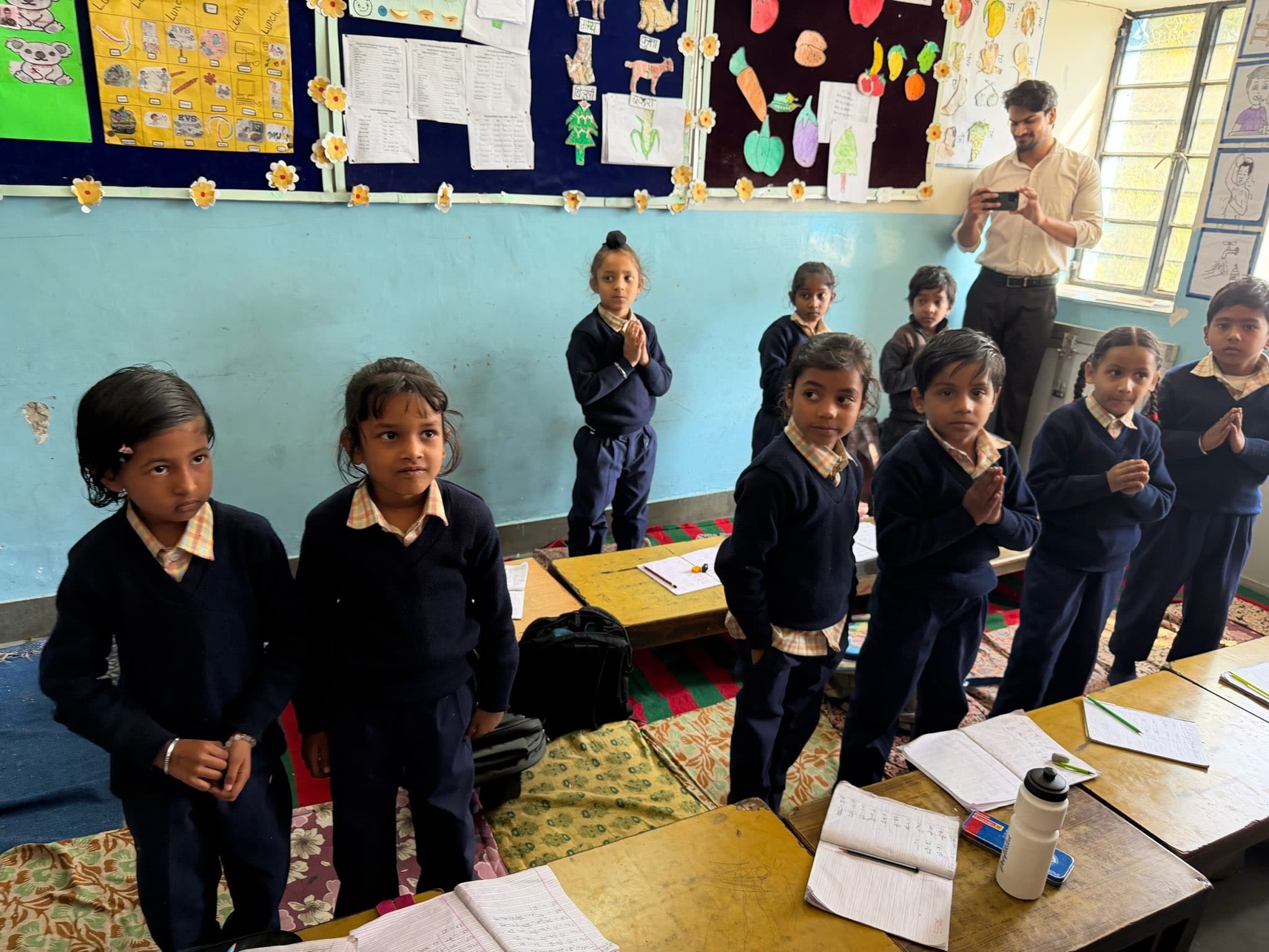 A row of young pupils stand to greet with folded hands, one girl smiling broadly, while their teacher looks on from the back of the classroom.