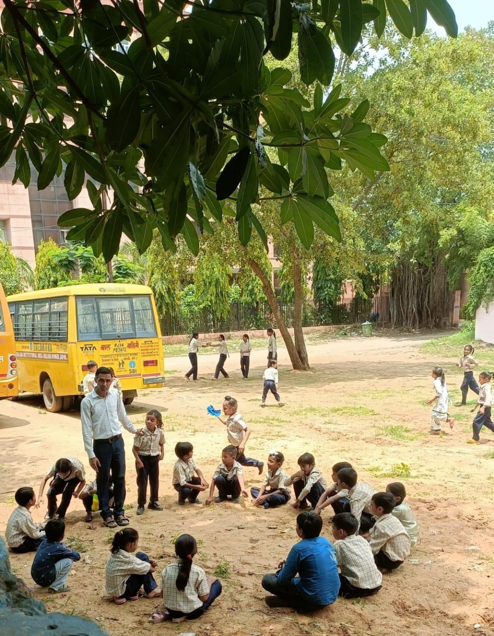 Children sit in a circle playing a game in the shaded schoolyard while a teacher stands among them; the school's yellow bus is parked under the trees behind.