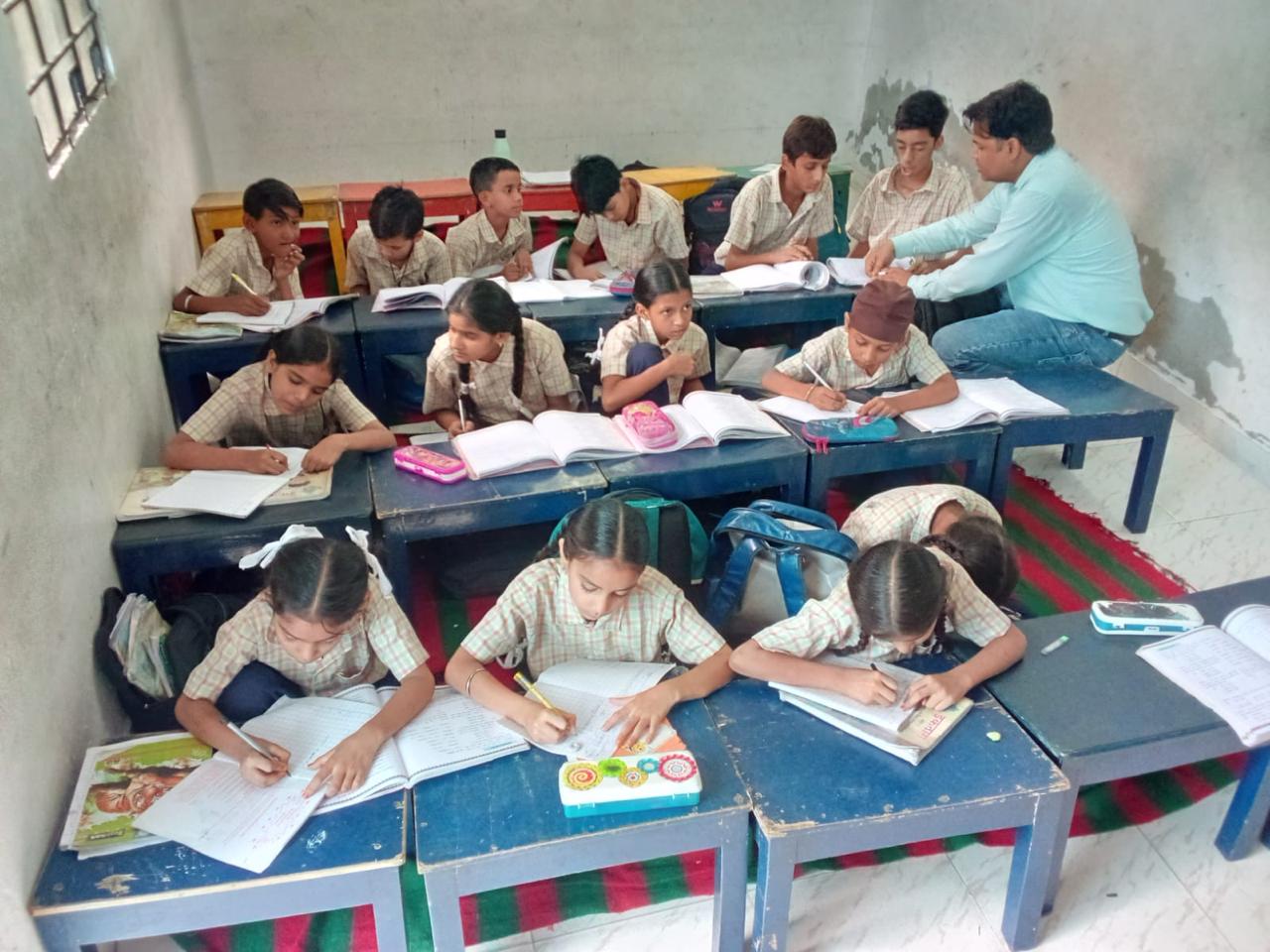 Older pupils write in their exercise books at low blue desks while their teacher leans in to help at the back of the classroom.