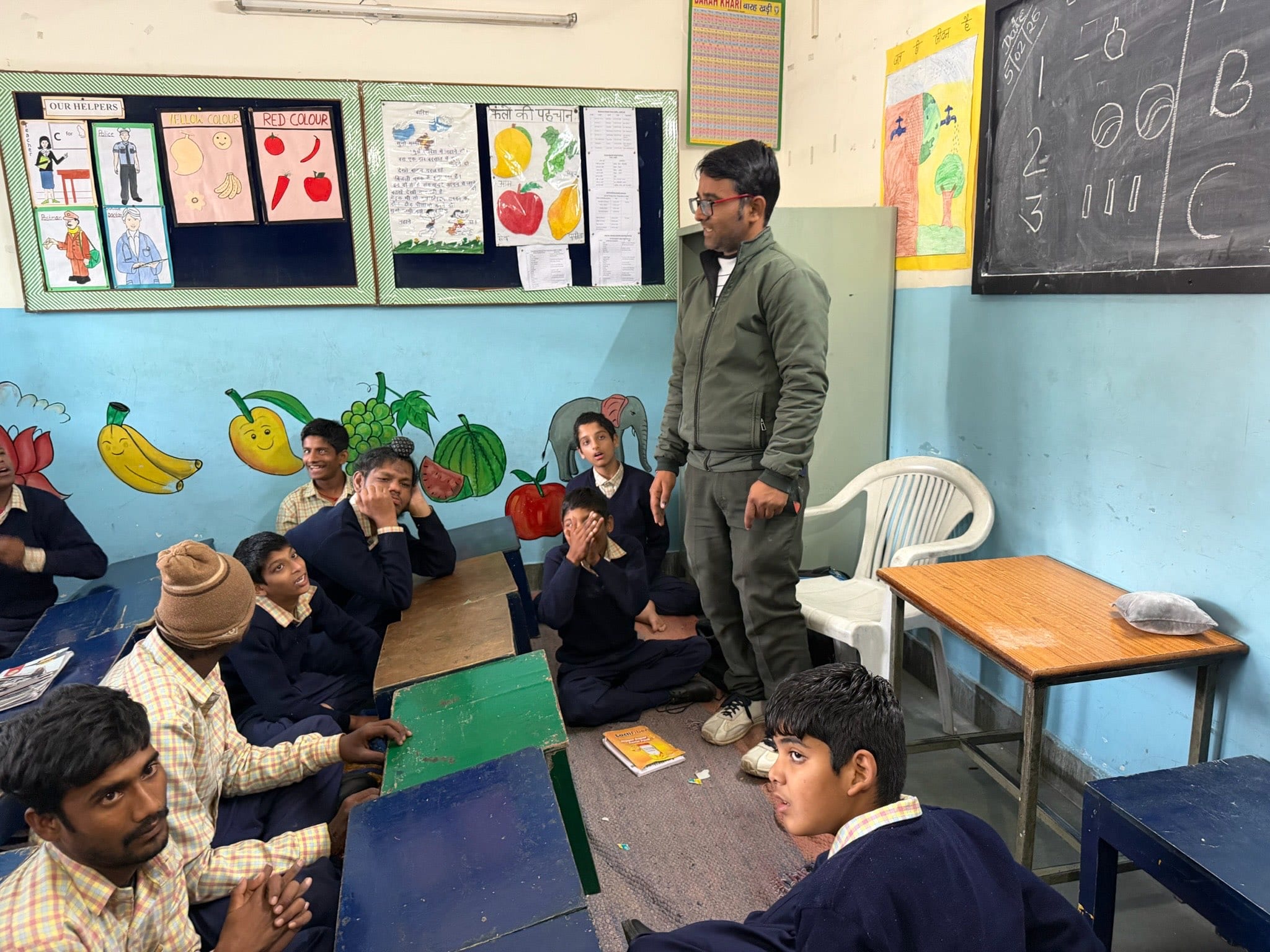 A young teacher stands smiling among his students in a classroom decorated with hand-drawn posters, a blackboard on the wall behind him.