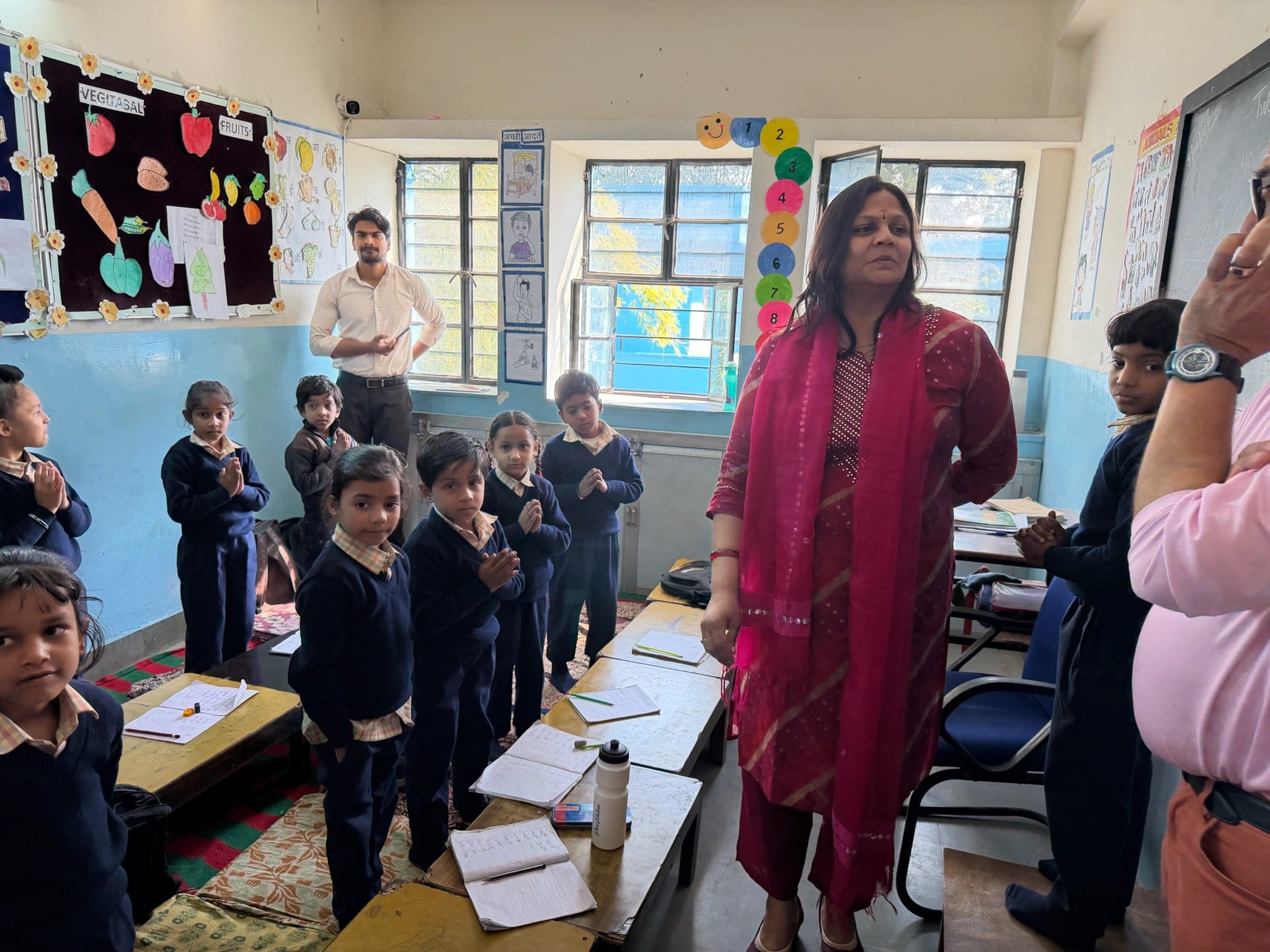 Children stand in greeting beside their desks as visitors are welcomed into the classroom.
